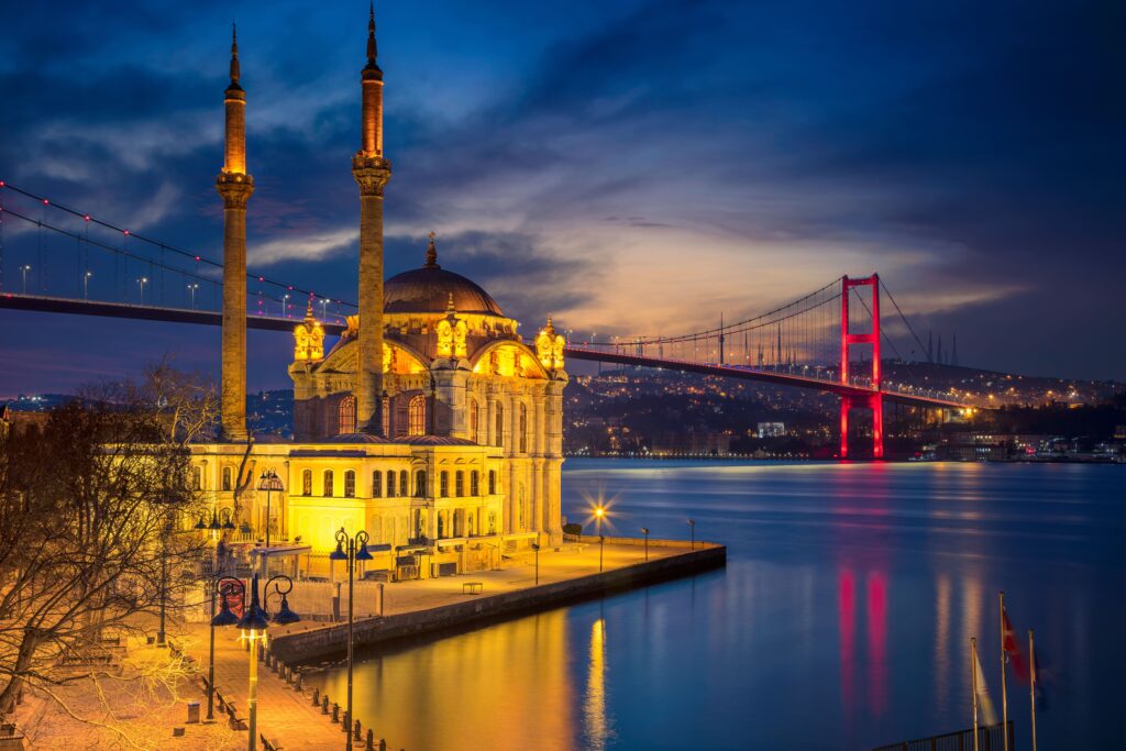 Die beleuchtete Moschee mit gelben Fassaden steht vor einer roten Brücke am Wasser