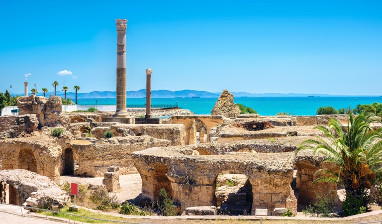 Ruinen einer antiken Stadt mit hellen Steinen, Palmen und türkisblauem Meer im Hintergrund