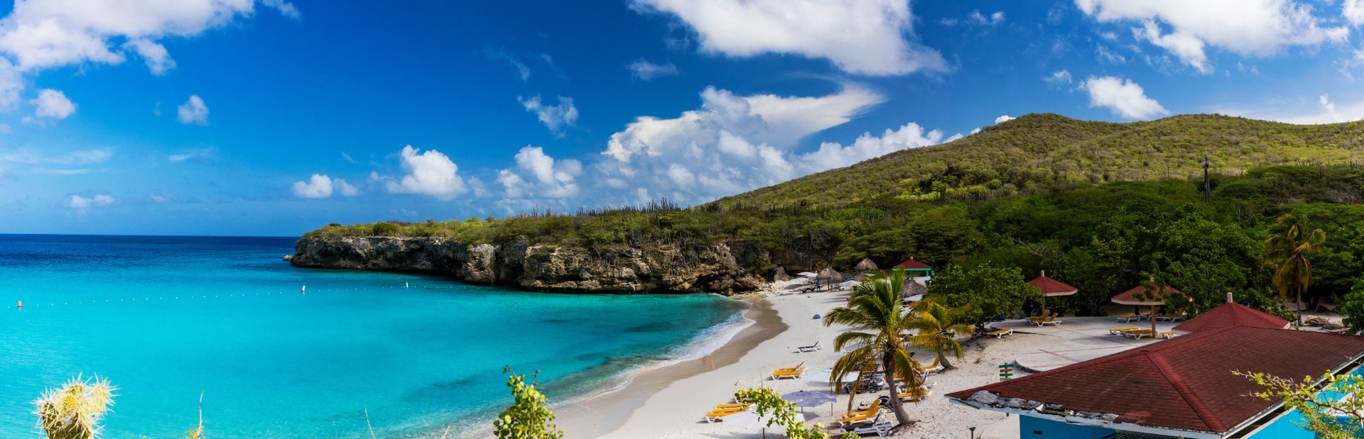 Ein tropischer Strand mit türkisblauem Wasser, weißen Sand und grünen Hügeln