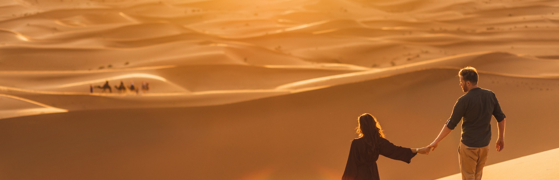 Ein Paar geht Händchen haltend durch golden beleuchtete Sanddünen bei Sonnenuntergang