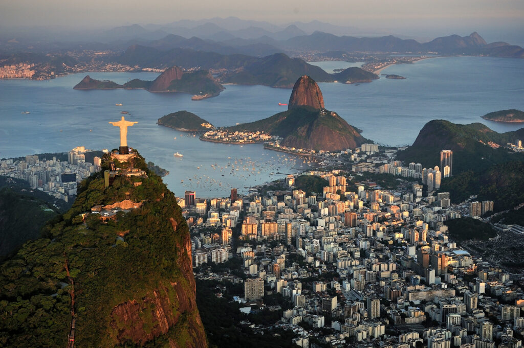Das Bild zeigt die Christusstatue in Rio de Janeiro vor einer sonnenbeleuchteten Stadt und grünen Hügeln