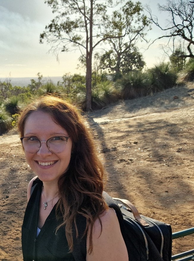 Eine Frau mit langen braunen Haaren lächelt freundlich im sonnigen Waldweg