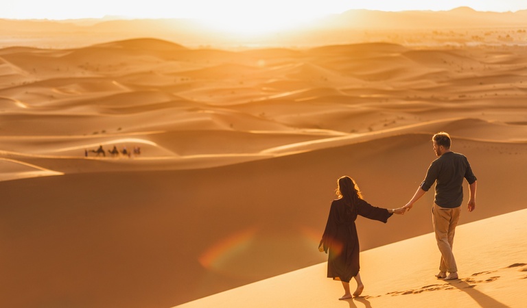 Ein Mann und eine Frau gehen Hand in Hand entspannt durch goldene Sanddünen