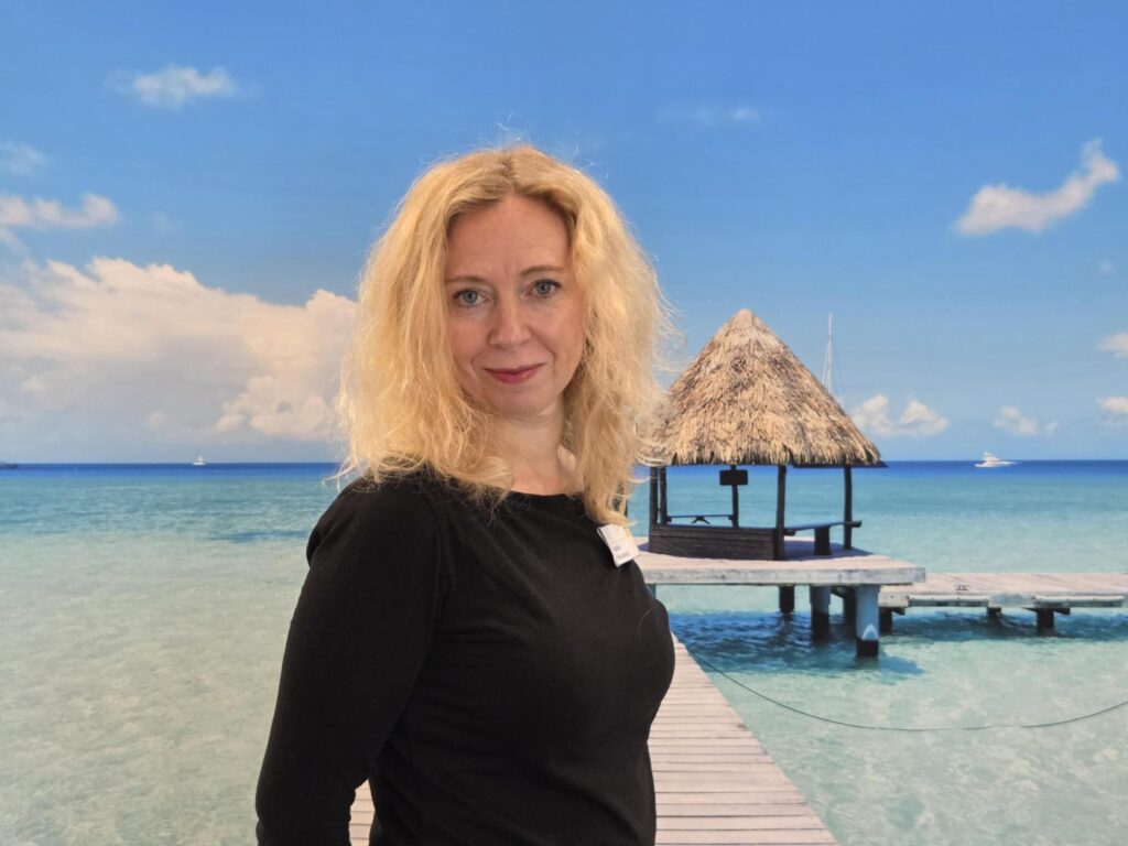 Eine Frau mit blondem lockigem Haar lächelt vor einer tropischen Strandkulisse