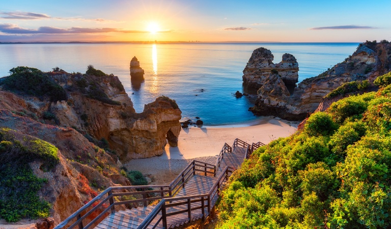 Eine Treppe führt zu einem sonnigen Strand mit goldenen Felsen und türkisfarbenem Wasser