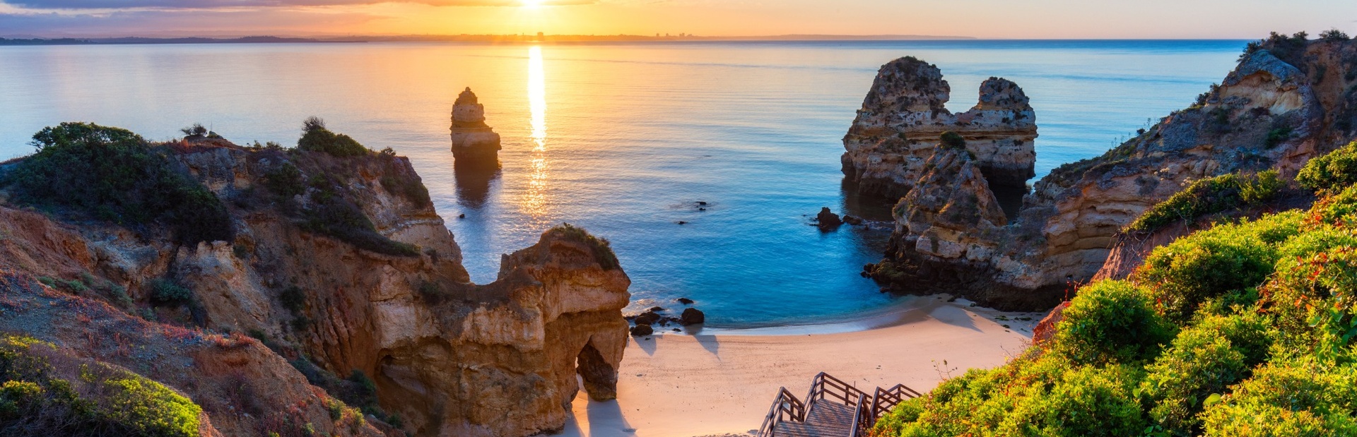 Klippen mit grüner Vegetation und hellem Sandstrand im Sonnenuntergang am Meer