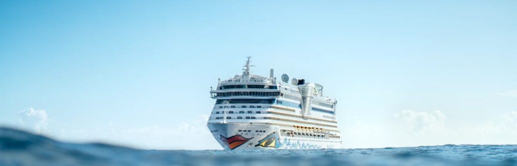 Ein großes, weißes Kreuzfahrtschiff mit rotem Mund lächelt fröhlich auf dem blauen Meer