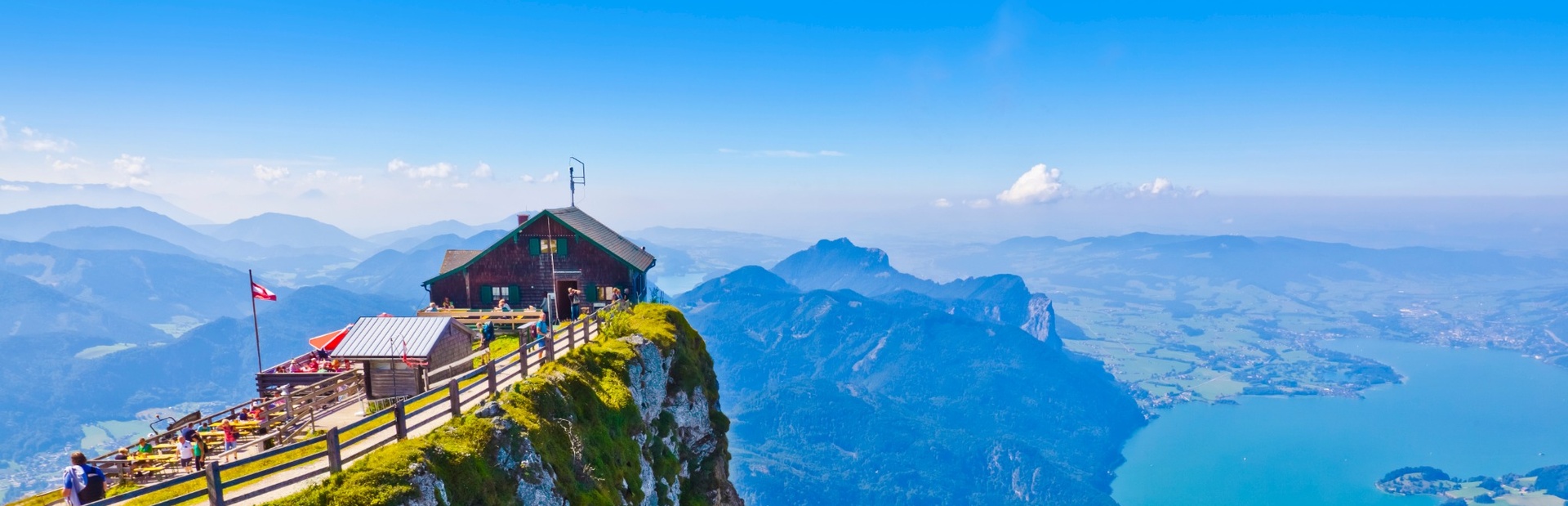 Eine Berghütte mit Personen entspannt auf einer sonnigen Wiese mit Blick auf See