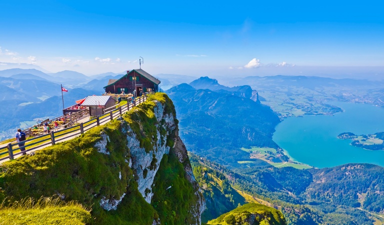 Ein Berghaus steht auf einer Klippe mit Blick auf türkisfarbenen See und Alpen
