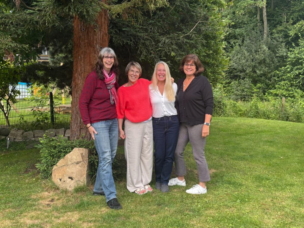 Vier lächelnde Frauen stehen in bunter Kleidung vor einem Baum im grünen Garten