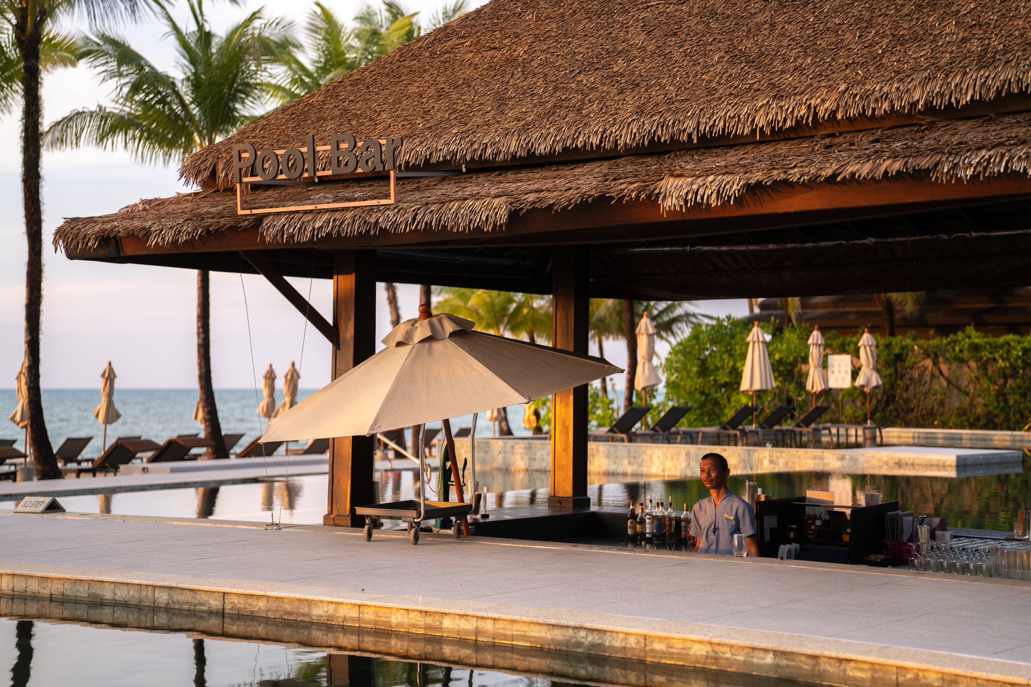 Ein Barkeeper mit neutralem Ausdruck steht hinter einer Bar am Pool unter Strohdach