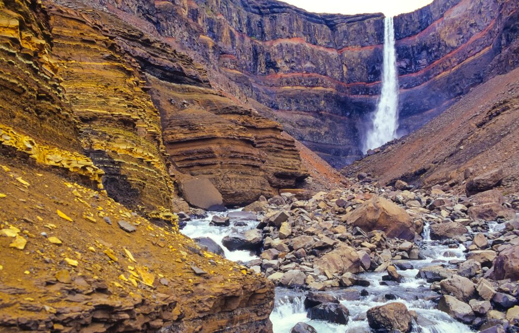 Ein hoher Wasserfall fließt zwischen bunt gestreiften Felsen über steinigen Fluss