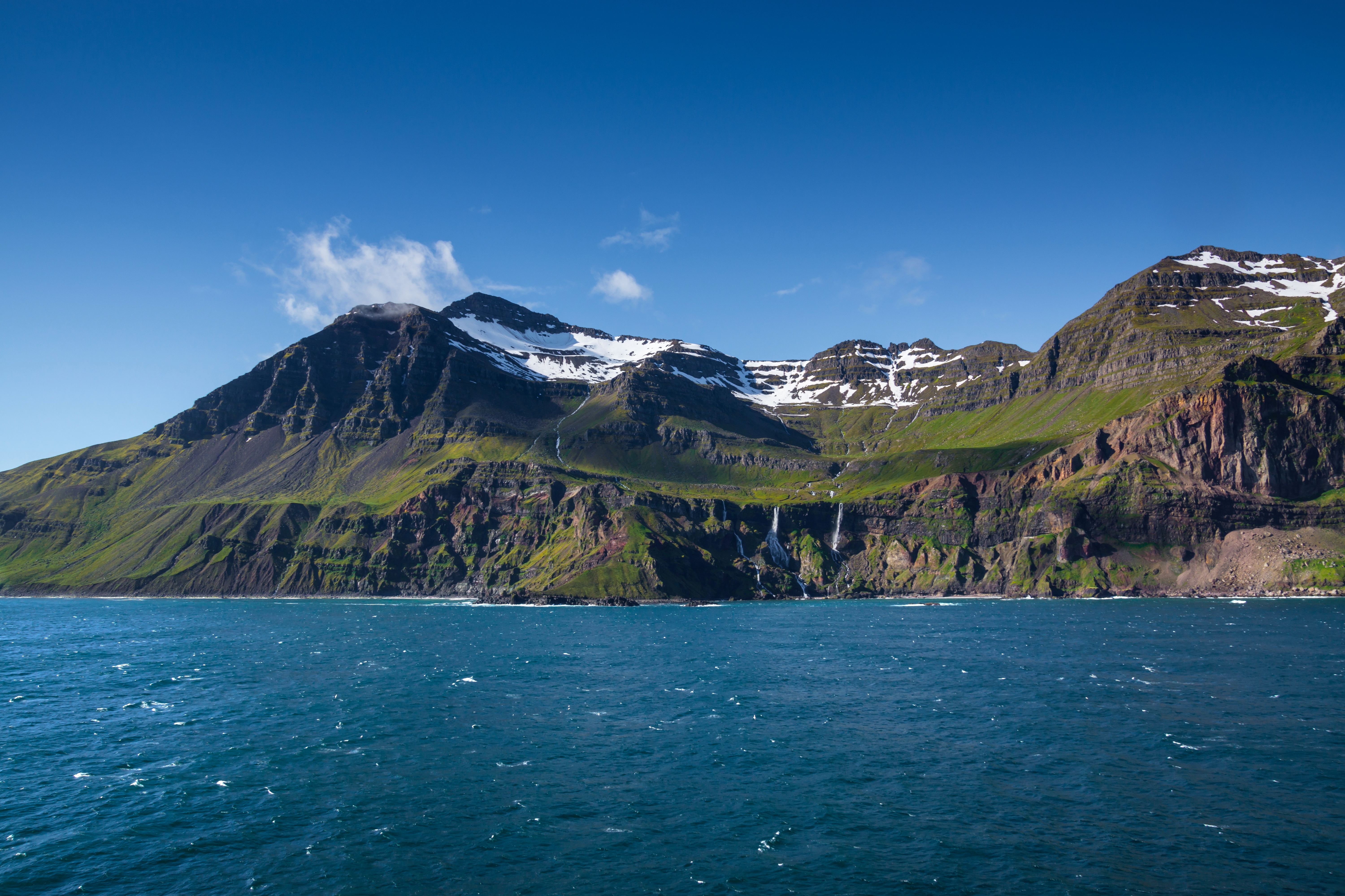 Klare blaue See mit grüner und brauner Berglandschaft, weißen Schneeflecken und Wasserfall