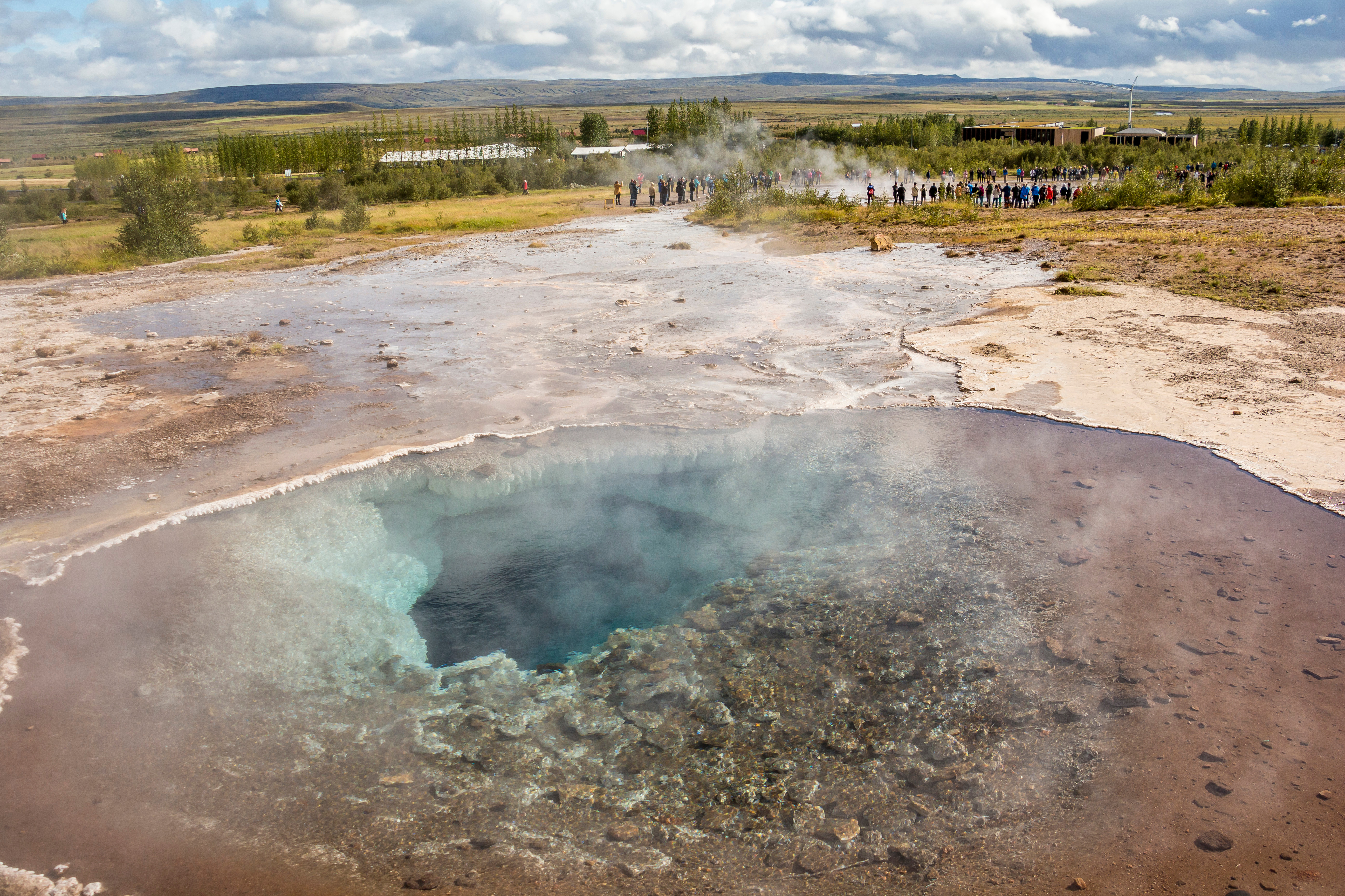 Eine Gruppe von Menschen betrachtet einen dampfenden blauen Geysir in einer weiten Landschaft