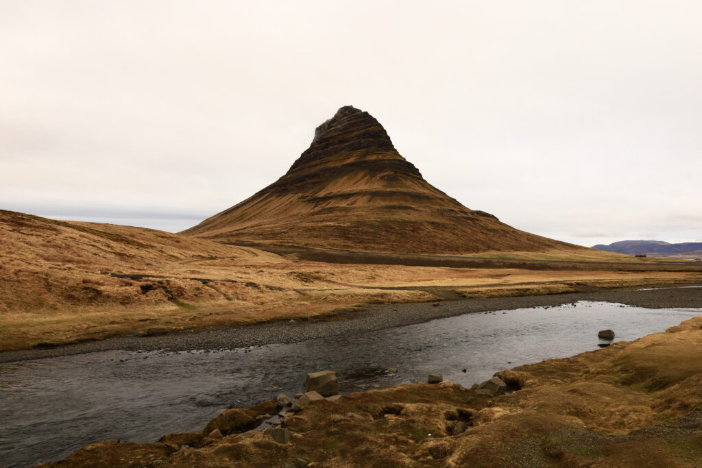 Ein brauner Berg mit markanter Spitze steht über einem ruhigen Fluss in weiter Landschaft