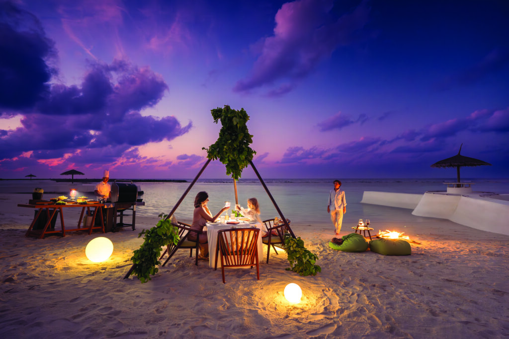 Zwei Personen sitzen entspannt am Tisch bei Abendessen am beleuchteten Strand