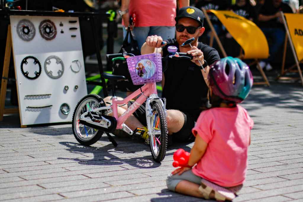 Ein Mann mit Sonnenbrille kniet neben einem rosa Kinderfahrrad, ein Kind mit Helm sitzt davor