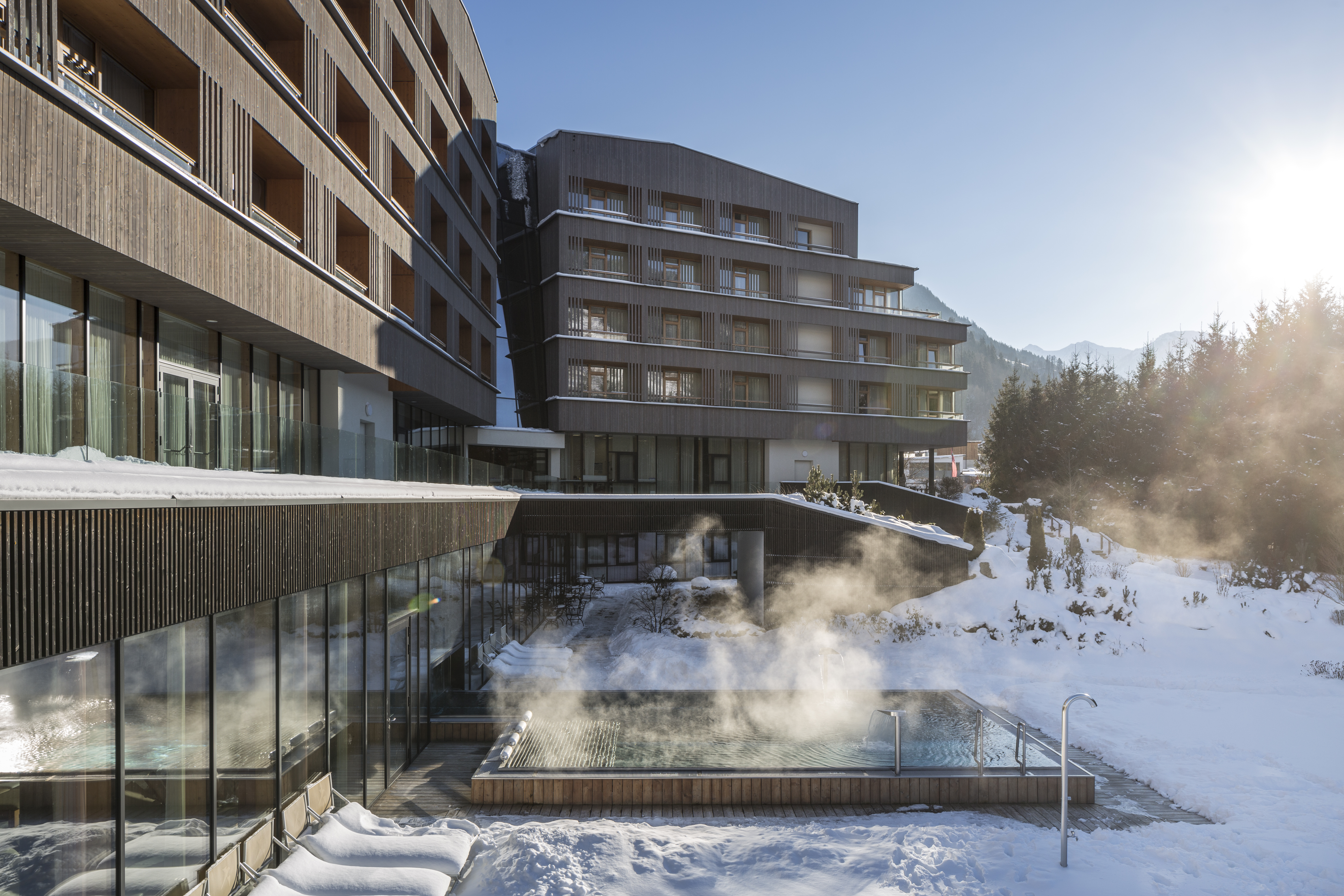 Modern Hotel mit Holzbalkonen im Winter, dampfender Außenpool und strahlende Sonne