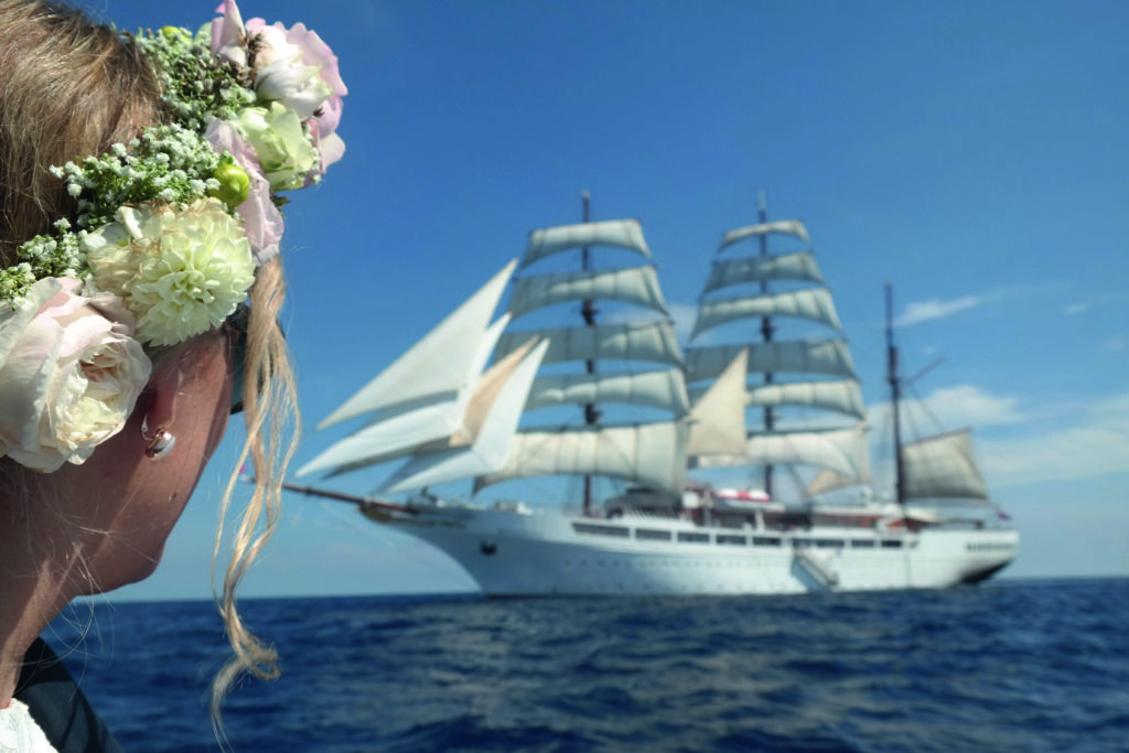 Eine blonde Person mit Blumenkranz schaut lächelnd auf ein großes weißes Segelschiff