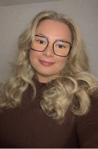 Eine Frau mit welligem, blondem Haar und Brille lächelt freundlich in einem braunen Oberteil