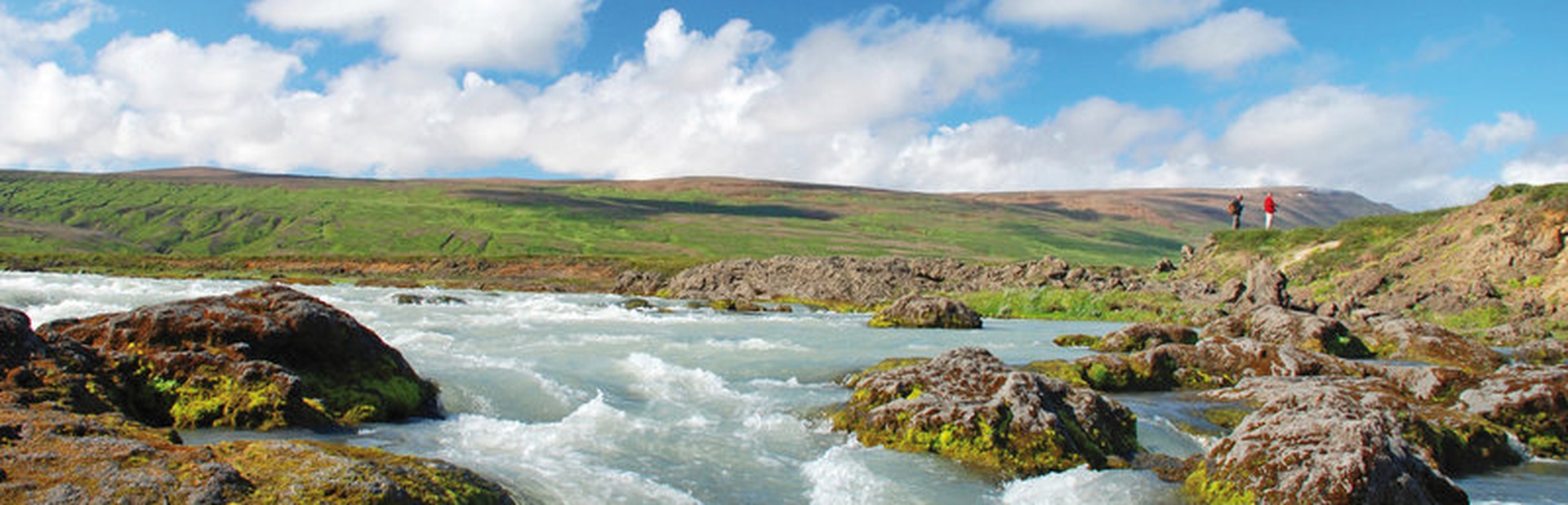 Zwei Personen stehen auf grünen Felsen am Fluss unter blauem Himmel mit Wolken