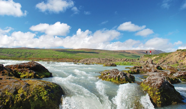 Zwei Personen stehen auf einem Felsen am Fluss, umgeben von grünem Moos und blauem Himmel