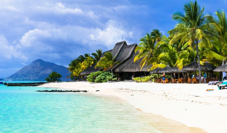 Ein tropischer Strand mit hellblauem Wasser, Palmen, Hütten und Menschen im Schatten