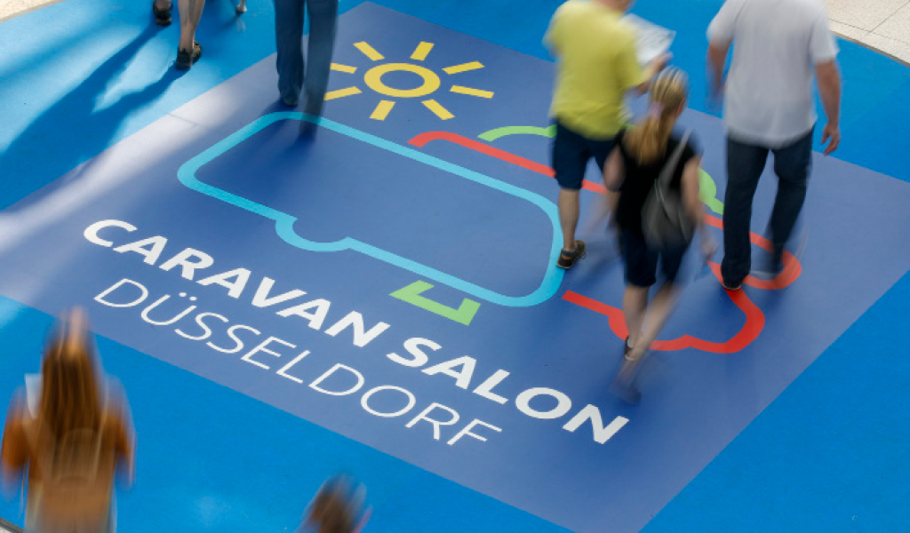 Mehrere Personen laufen auf einem blauen Messeboden mit buntem Caravan-Symbol