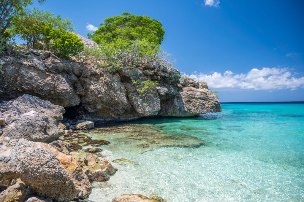 Ein sonniger Strand mit klar türkisfarbenem Wasser und felsigen Klippen, grüne Bäume