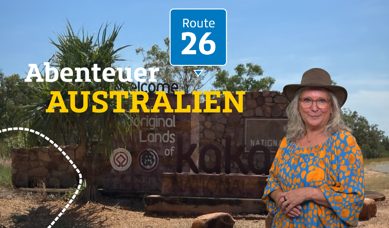 Eine freundlich lächelnde Frau mit Brille und Hut steht vor dem Kakadu Nationalpark-Schild